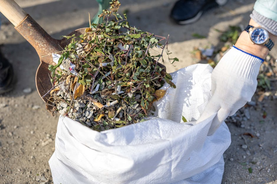 A person wearing a white glove and a wristwatch is holding a large white sack filled with a mix of garden waste and small debris, including twigs, leaves, and gravel. The scene appears to take place outdoors on a paved or gravel surface, with a metal shovel resting on top of the collected waste, partially visible in the upper left corner. The background includes a patch of green foliage and some scattered small items on the ground. The image relates to waste collection and disposal services, showcasing the process of gathering garden or outdoor rubbish, which might be handled by private waste removal providers like Waste Disposal Streatham, as part of alternative waste handling options.