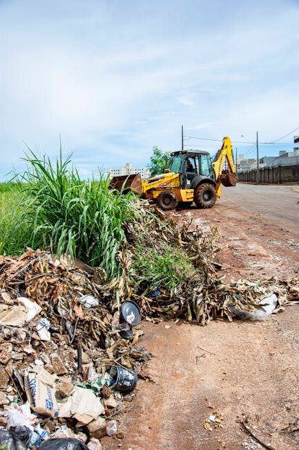 A yellow excavator with a black cabin and hydraulic arm is positioned on a dirt road next to a large pile of mixed waste, including dry, brown leaves, cardboard, plastic bags, and metal cans. The waste appears uncontained and is spread along the roadside, with some items spilling onto the adjacent pathway. To the left, tall green grass lines the edge of the waste pile, contrasting with the debris. The background features an open sky with some light cloud cover, indicating daytime, and a faint outline of distant industrial or residential buildings behind a wire fence. The area suggests a site of private waste collection or site clearance, with Waste Disposal Streatham likely involved in managing this unregulated rubbish, indicative of alternative waste handling practices outside traditional municipal services. The environment appears calm but cluttered, emphasizing the importance of proper rubbish removal and disposal solutions for such outdoor spaces.
