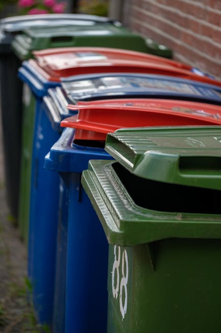 A row of large outdoor wheelie bins lined up along a sidewalk next to a brick wall, with the bins primarily blue, green, and red in colour. The green bin in the foreground has a ridged lid and a partially visible recycling symbol on its side. Behind it, the blue bins have smooth lids, with some showing slight openings or angles. The red lids are positioned on top of the corresponding bins, with their edges slightly overhanging the sides. The bins are placed on a paved area adjacent to a residential building, suggesting an arrangement for waste collection or private waste disposal. The environment appears clean, with the bins neatly aligned, indicating scheduled rubbish or recyclables disposal service. This scene reflects typical private waste collection practices, possibly undertaken by Waste Disposal Streatham, to manage waste outside residential premises efficiently and securely, supporting the context of alternative rubbish collection methods near Streatham Common.